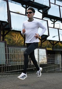 Homem em boa forma, vestindo uma camisa de manga longa branca e leggings pretas, usando óculos de sol e tênis esportivos brancos, correndo numa superfície pavimentada.