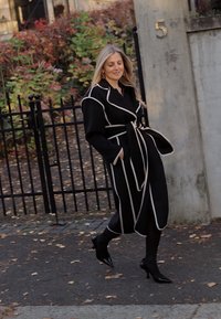 Black coat with white piping, belted waist, long sleeves, and flowing design. Worn with fitted black pants and pointed black heels.
