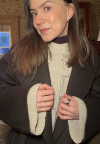 Γυναίκα με μακριά καστανά μαλλιά που φοράει σκούρο παλτό πάνω από εκρού πλεκτό πουλόβερ, κρατώντας τα πέτα του παλτού με τα δύο χέρια μέσα σε κτήριο κοντά σε παράθυρο.