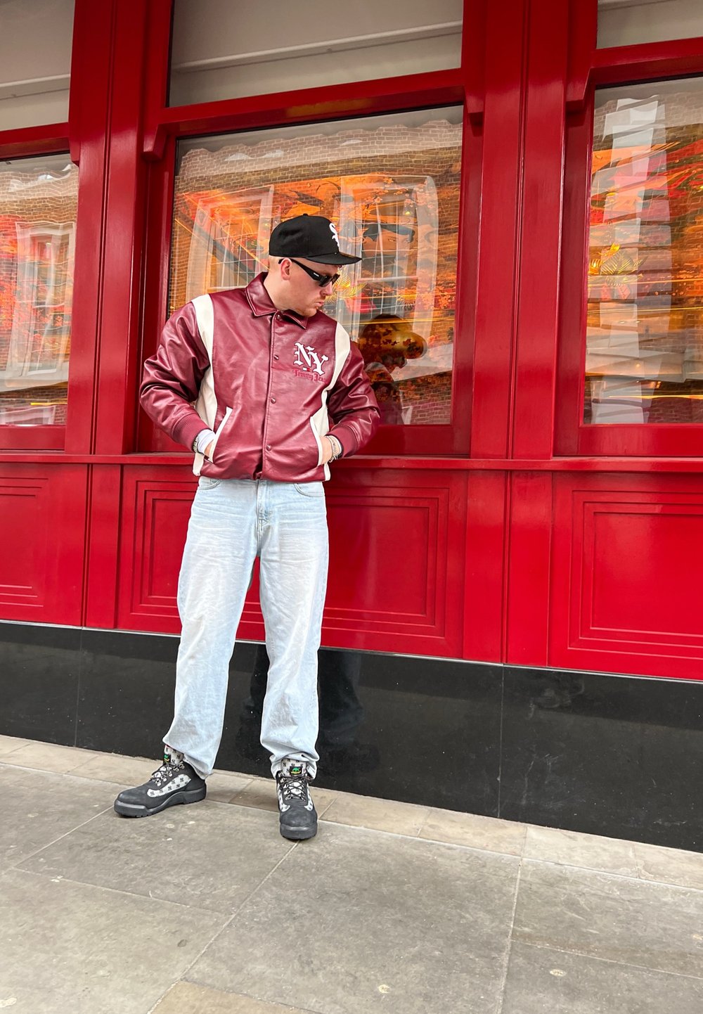 Giacca in pelle rossa e bianca con logo "NY", pantaloni azzurri chiari e stivali neri resistenti con calzini a fantasia, in piedi davanti a uno sfondo di finestra rossa.