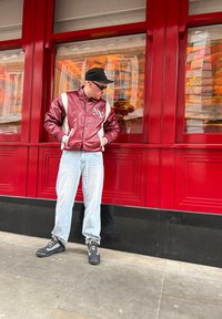 Giacca in pelle rossa e bianca con logo "NY", pantaloni azzurri chiari e stivali neri resistenti con calzini a fantasia, in piedi davanti a uno sfondo di finestra rossa.