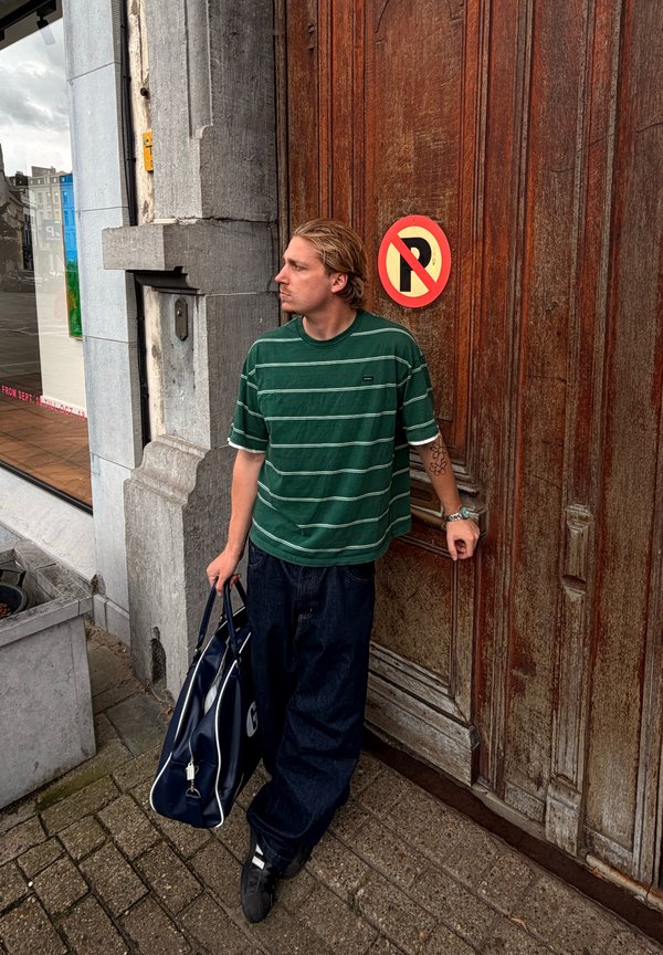 Pessoa com uma camisa às riscas verdes e calças escuras encostada a uma porta de madeira com uma placa de proibição de estacionamento, segurando uma mala azul numa calçada da cidade.