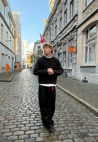 Suéter de punto negro sobre una camisa blanca, combinado con jeans negros holgados. Callejón empedrado con edificios históricos y una bandera roja.