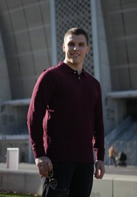 Maroon long-sleeve polo shirt with a collar and button detail, paired with dark jeans. Person holding sunglasses, standing outdoors.