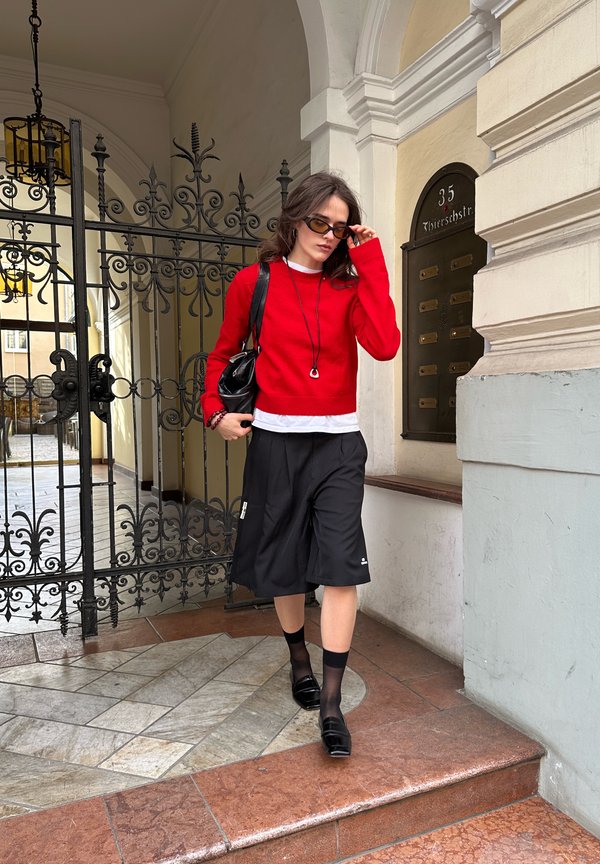 Femme en pull rouge, jupe noire et lunettes de soleil descendant des marches en pierre près d'une porte en fer forgé décorative et d'une façade de bâtiment beige.