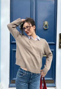 Béžový, skrátený sveter s výstrihom do V a rebrovaným lemom, kombinovaný s bielou košou s golierom a modrými džínami. Drží červenú kabelku. Pozadie modrých dverí.