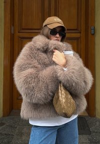 Barna műszőrméből készült dzseki széles gallérral, fehér inggel és világoskék farmerrel párosítva. Egy kávésbögrét és egy fonott aranyszínű kézitáskát tart a kezében.