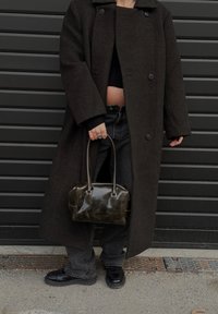 Manteau en laine foncée avec de grands boutons, pull noir court, jean taille haute, chaussures noires brillantes et sac à main sombre et brillant.