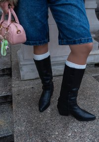 Μαύρες δερμάτινες μπότες μέχρι το γόνατο με μυτερές μύτες και τετράγωνα τακούνια, σ�υνδυασμένες με λευκές ριμπέ σόκες και φορεμένες με τζιν σορτς.