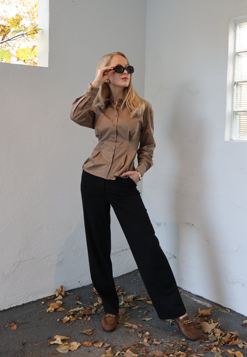 Hnedá košeľa na gombíky s nafúknutými rukávmi, čierne široké nohavice a hnedé topánky s ozdobami. Modelka v čiernych slnečných okuliaroch pózuje v interiéri.