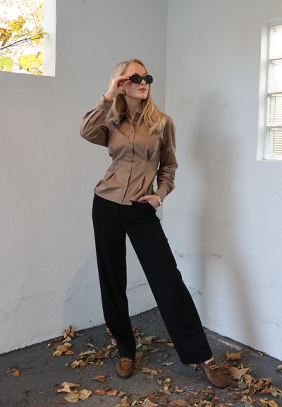 Camisa marrón abotonada con mangas abullonadas, pantalones negros de pierna ancha y zapatos marrones con detalles. Modelo con gafas de sol negras posando en el interior.