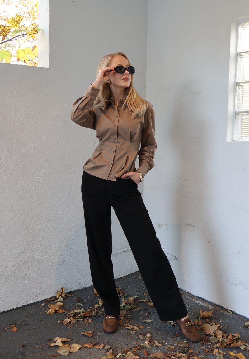 Brun skjorte med knapper og ballonærmer, sorte bukser med vide ben og brune sko med detaljer. Model iført sorte solbriller poserer indendørs.