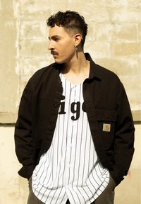 Giacca nera a bottoni sopra una maglietta da baseball bianca a righe con scritte nere. Capelli corti e orecchino visibili.