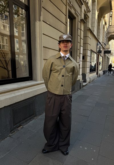 Chaqueta corta verde oliva claro con botones negros, combinada con pantalones oscuros oversized que tienen un detalle de cadena, y calzada con zapatos negros pulidos.