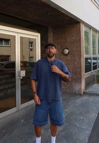 Mand iført en blå kasket, briller, en oversized blå T-shirt, denimshorts, hvide sokker og sandaler står udenfor en bygning med glasdøre.