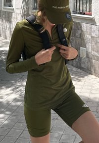 Camisa de manga larga verde y pantalones cortos, ambos con el logo "The North Face". Una gorra a juego completa el atuendo atlético, llevado con una mochila negra.