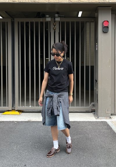 Camiseta negra de manga corta con el texto "Carhartt", pantalones cortos de mezclilla azul claro, camisa de cuadros gris atada a la cintura, zapatos marrones y calcetines blancos.