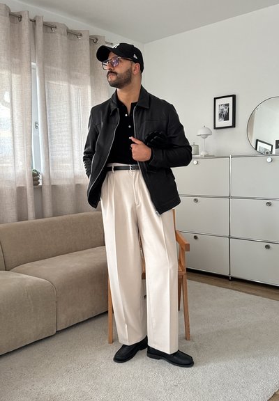 Chaqueta de piel negra, camiseta polo negra, pantalones de pierna ancha color crema, zapatos negros, gafas y gorra; de pie en una sala de estar con decoración moderna.