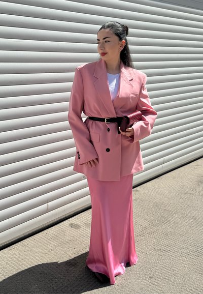Blazer rosa con dos botones sobre una falda de satén a juego. Combinado con un cinturón negro y un pequeño bolso burdeos. Textura suave, diseño formal.