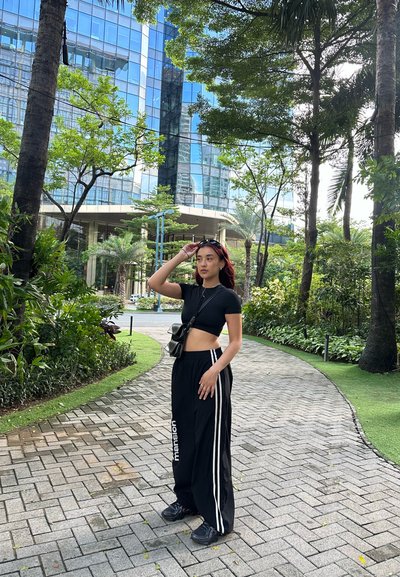 Camiseta corta negra y pantalones de pierna ancha negros con rayas blancas; pequeña bolsa negra; el entorno incluye vegetación y un edificio moderno de cristal.