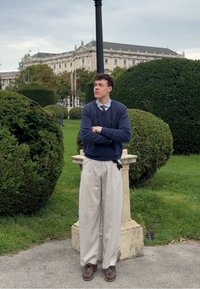 Una persona si trova vicino a cespugli potati, indossando un maglione blu navy, pantaloni beige chiaro e scarpe marroni. L'ambiente include un pilastro di pietra e vegetazione.