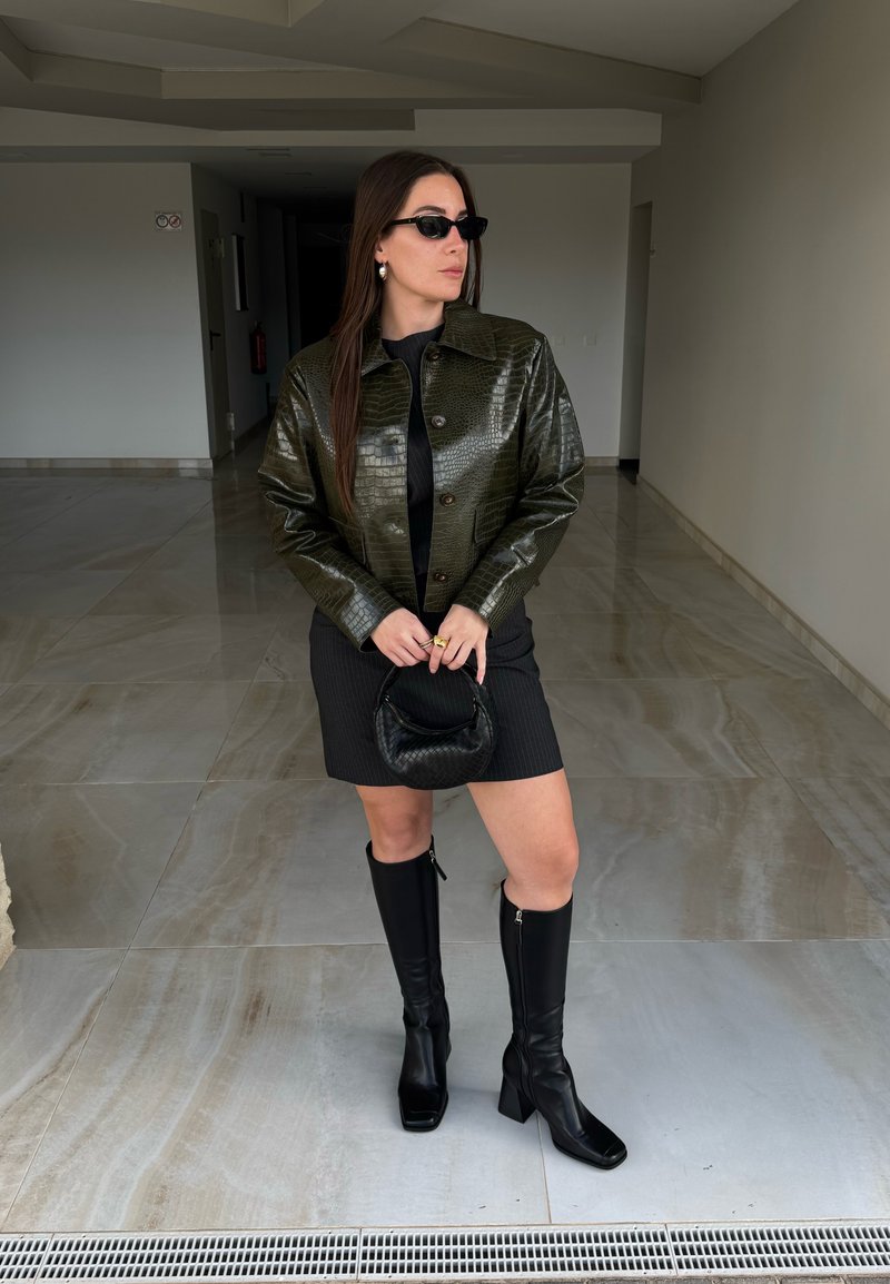 Femme portant une veste texturée vert foncé, une mini-robe noire, des bottes noires montant jusqu'aux genoux, des lunettes de soleil, tenant un petit sac à main noir dans un couloir carrelé.