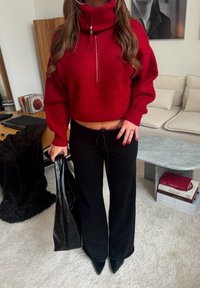 Femme portant un pull rouge court et un pantalon noir, tenant un sac en cuir noir, debout dans un salon moderne avec des livres et un canapé.