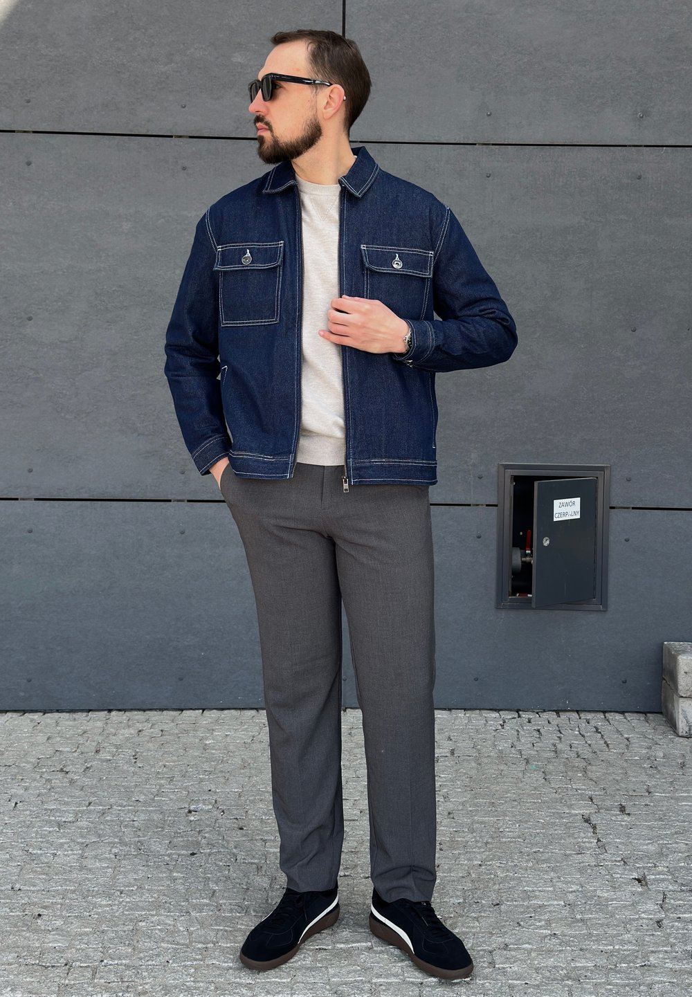 Chaqueta de mezclilla azul oscuro con dos bolsillos en el pecho, suéter gris debajo, pantalones grises y zapatillas negras con acentos blancos. Fondo de concreto.