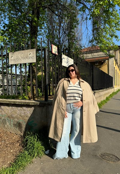 Gabardina beige de gran tamaño, suéter a rayas, jeans de pierna ancha de color azul claro y gafas de sol negras, de pie cerca de una valla con señales de tráfico.