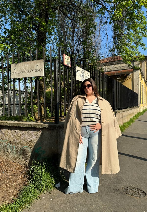 Trench coat beige oversize, maglione a righe, jeans a gamba larga di un blu chiaro e occhiali da sole neri, in piedi vicino a una recinzione con segnali stradali.