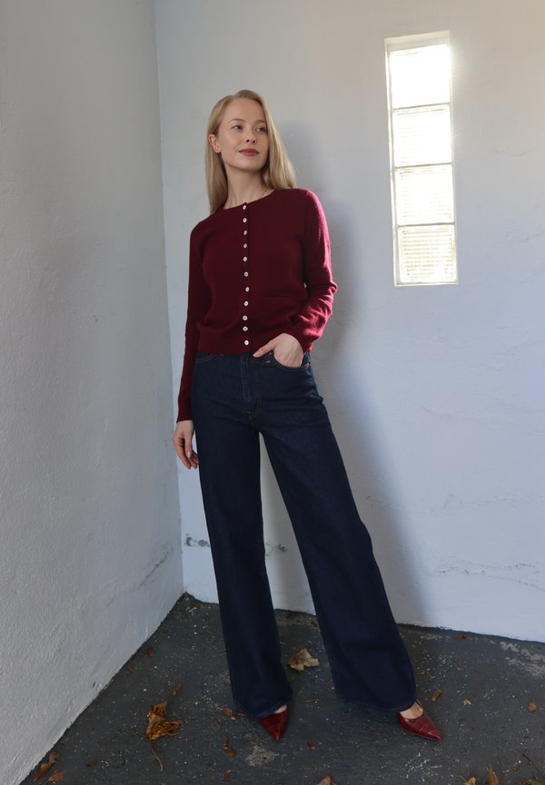 Cárdigan de punto color granate con cierre de botones, jeans azules de cintura alta y tacones rojos con punta puntiaguda. Pared de color gris claro con una ventana rectangular.