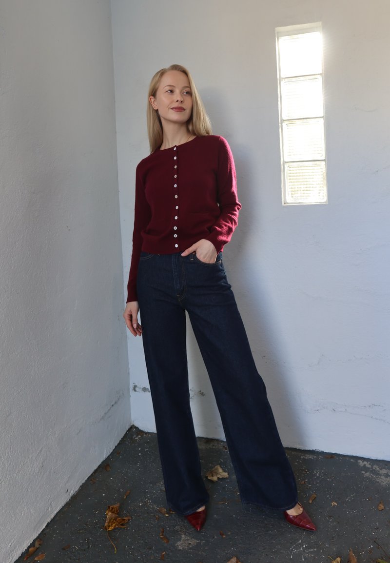 Bordeauxrød strikket cardigan med knaplukning, blå højttaljede jeans og røde spidse hæle. Lys grå væg med et rektangulært vindue.