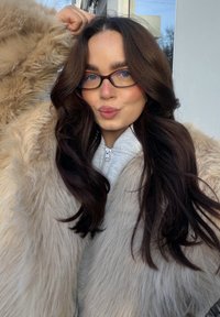 Długie, falowane ciemne włosy, ubrana w jasnoszary bluzy z zamkiem i beżowy płaszcz z imitacji futra, stoi na tle jasnego tła. Okulary z niebieskimi akcentami.