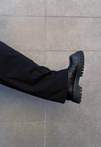 Bottine noire en cuir avec une semelle épaisse et crantée, associée à un pantalon noir. La chaussure présente des coutures minimalistes et une finition lisse.
