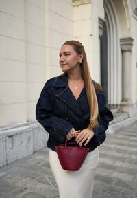 Jachetă din denim cu guler larg, mâneci lungi și accesorii din aur, purtată cu o fustă crem ajustată și o geantă mică burgundă.