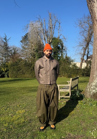 Chaqueta bomber marrón, pantalones holgados verdes y gorro naranja. La chaqueta tiene dos bolsillos frontales; los pantalones cuentan con una cintura con cordón y puños anchos.