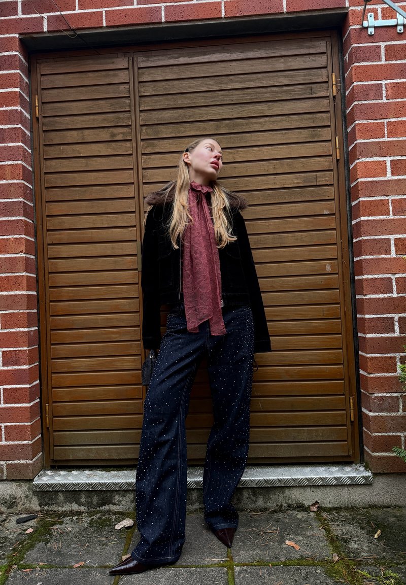 Veste en velours noir avec col en fourrure, écharpe bordeaux, pantalon en denim foncé avec ornements, et talons pointus devant un fond en bois.