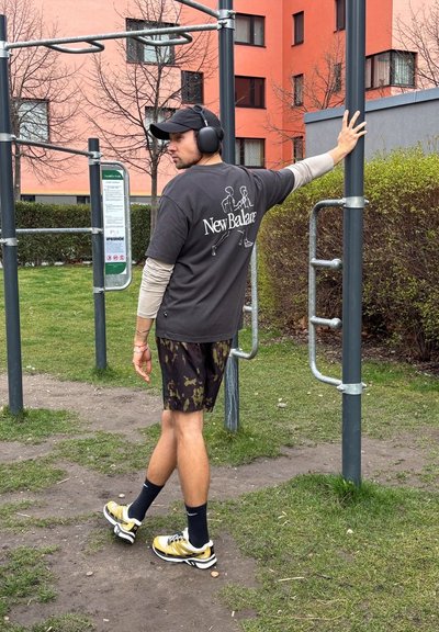 Atleta con una camiseta negra de New Balance, pantalones cortos de camuflaje y zapatillas doradas/blancas, agarrándose a una barra de ejercicios en un entorno de parque.