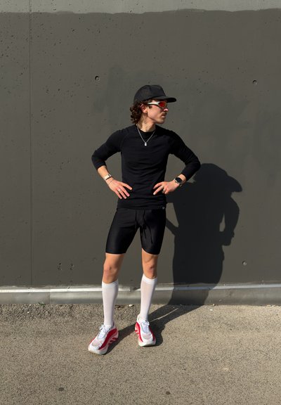 Persona con ropa deportiva negra, calcetas blancas hasta la rodilla, zapatillas deportivas rojas y blancas, gafas de sol y gorra, de pie con las manos en las caderas frente a una pared gris.