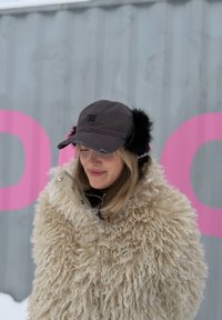 Femme portant un grand manteau crème duveteux et une casquette noire avec des cache-oreilles, debout à l'extérieur devant un mur gris avec des lettres roses.