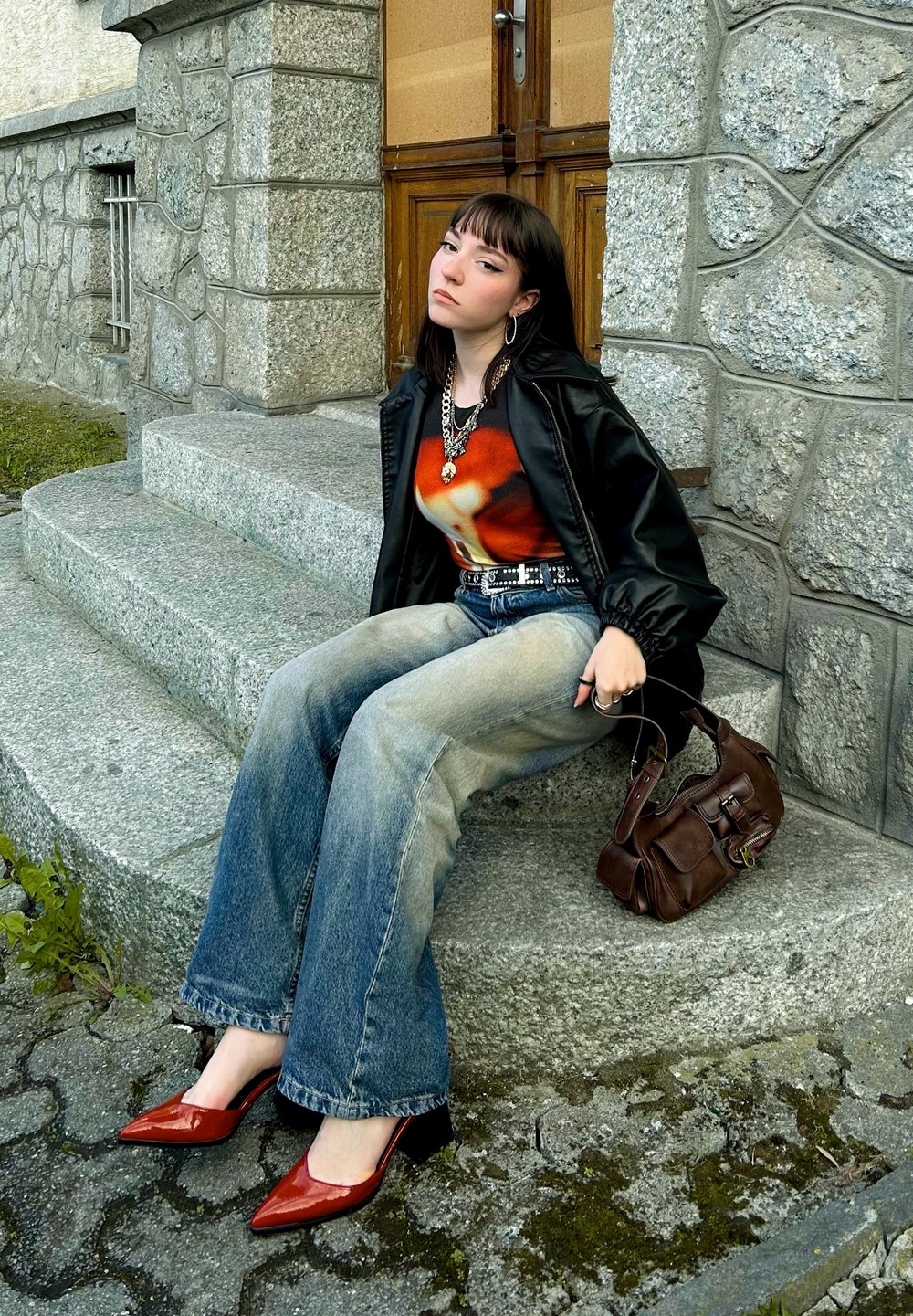 Μαύρο κοντό σακάκι, τοπ με κόκκινες και πορτοκαλί διακοσμήσεις, ανοιχτό μπλε καμπάνα τζιν, κόκκινες μυτερές γόβες και καφέ τσάντα σε γκρίζες πέτρινες σκάλες.