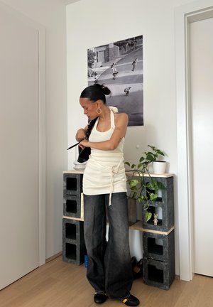 Top blanco sin mangas con un detalle de lazo en la parte de atrás, combinado con jeans de mezclilla anchos y oscuros. Bolsa negra, planta de interior y un cartel en la pared al fondo.