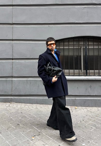 Abrigo de lana negra, camisa azul, pantalones anchos negros y gafas de sol marrones de gran tamaño. Lleva un bolso de cuero negro. Fondo de pared gris.