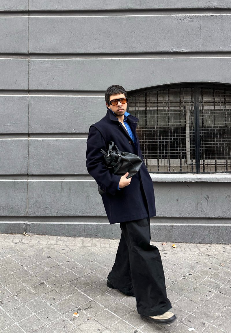 Manteau en laine noire, chemise bleue, pantalon noir large et lunettes de soleil marron surdimensionnées. Porte un sac en cuir noir. Fond mur gris.