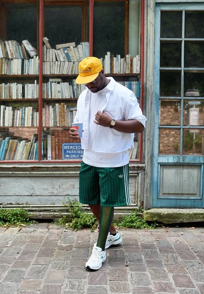 Pantalones cortos a rayas verdes, camisa blanca, gorra amarilla y zapatillas blancas. Lleva un reloj y sostiene una taza. El fondo presenta estanterías de libros.