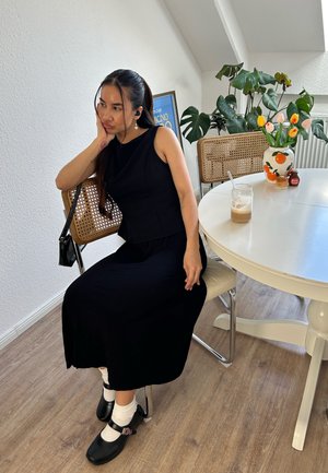 Vestido negro ajustado sin mangas con un detalle de peplum, combinado con calcetas blancas y zapatos de charol negros estilo Mary Jane, sentada en una mesa redonda blanca.