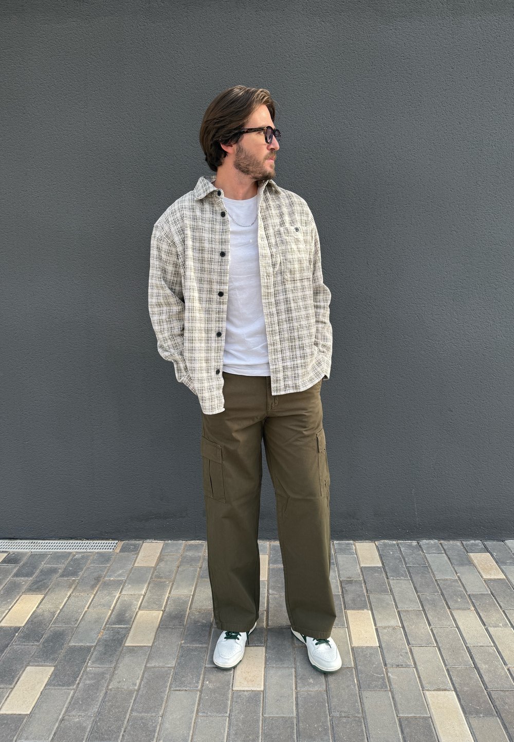 Chemise beige à carreaux avec boutons noirs, portée sur un t-shirt blanc. Pantalon cargo vert olive, baskets blanches avec des accents verts, debout sur des pavés.