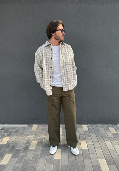Camisa de cuadros beige con botones negros, superpuesta sobre una camiseta blanca. Pantalones cargo verde oliva, zapatillas blancas con detalles verdes, de pie sobre pavimento.
