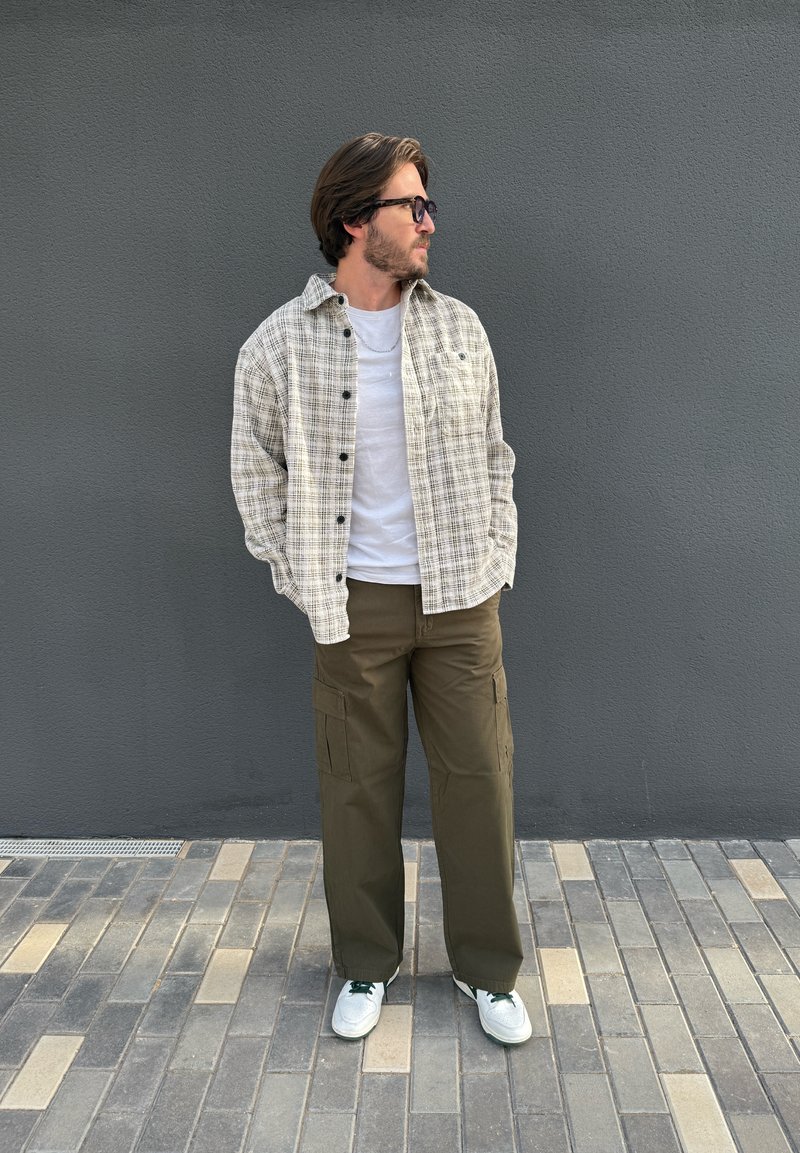 Chemise beige à carreaux avec boutons noirs, portée sur un t-shirt blanc. Pantalon cargo vert olive, baskets blanches avec des accents verts, debout sur des pavés.