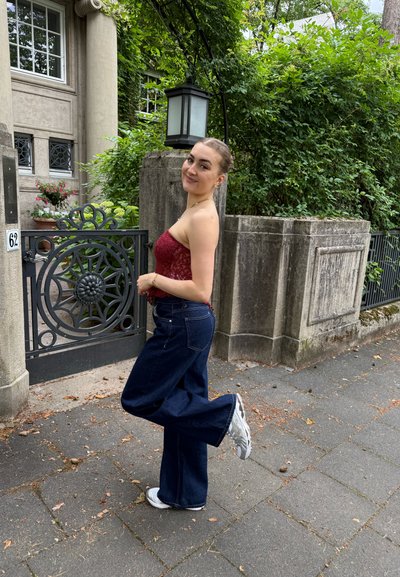 Top de encaje rojo con un dobladillo con volantes, pantalones de mezclilla oscuros acampanados y zapatillas blancas. De pie junto a una puerta decorativa y una vegetación exuberante.